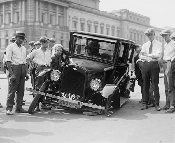 A group of young people gather around a broken vehicle in the early 20th century